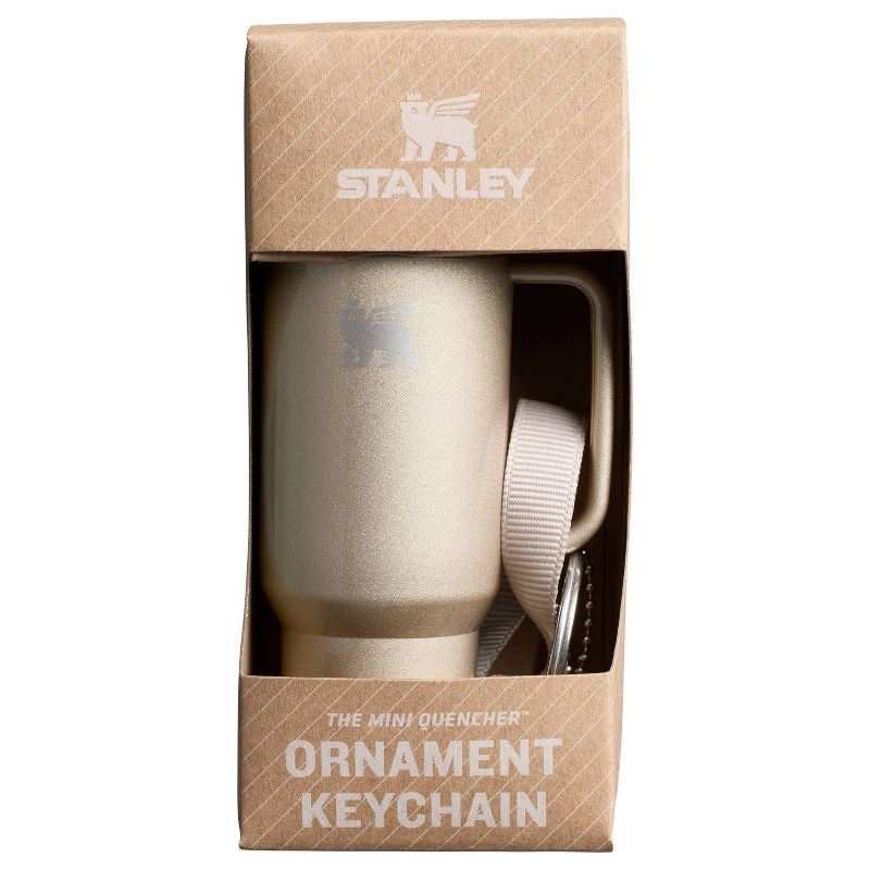 A mini Stanley tumbler-shaped keychain with a beige finish is displayed in packaging, alongside a white strap for attachment.
