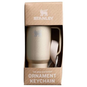 A mini Stanley tumbler-shaped keychain with a beige finish is displayed in packaging, alongside a white strap for attachment.