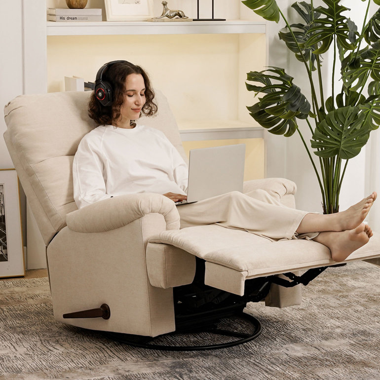 A person is sitting in a beige recliner chair with the footrest extended, using a laptop, and wearing headphones. There is a large green plant beside the chair.