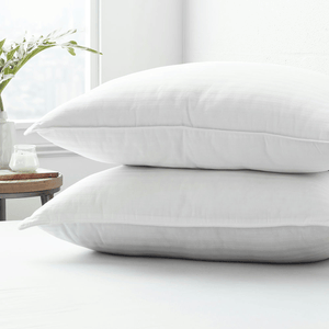 Two fluffy white pillows stacked on a bed, with a small table beside them holding a candle and a vase with green leaves.