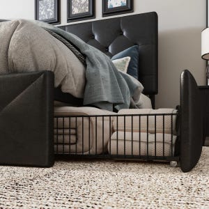 Black leather headboard, tufted design, featuring a wooden bed frame with an under-bed storage basket on wheels holding folded blankets. Beige and blue pillows and throws are arranged on the bed over a textured carpet.