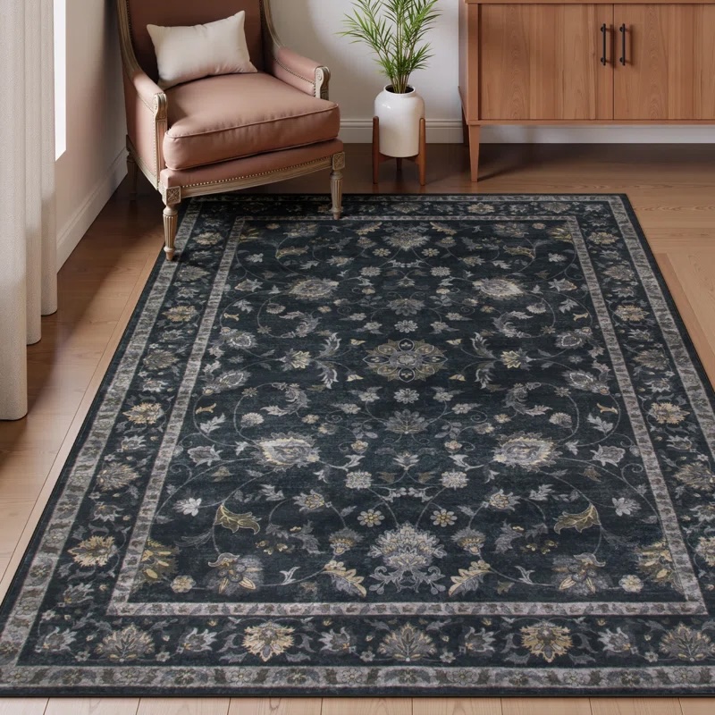 A dark floral-patterned area rug placed in a room with a wooden floor, accompanied by an upholstered chair, small pillow, indoor plant, and a wooden cabinet.
