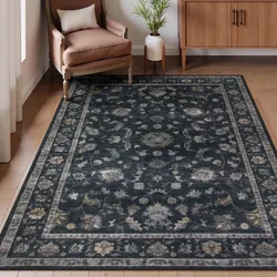 A dark floral-patterned area rug placed in a room with a wooden floor, accompanied by an upholstered chair, small pillow, indoor plant, and a wooden cabinet.