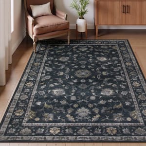 A dark floral-patterned area rug placed in a room with a wooden floor, accompanied by an upholstered chair, small pillow, indoor plant, and a wooden cabinet.
