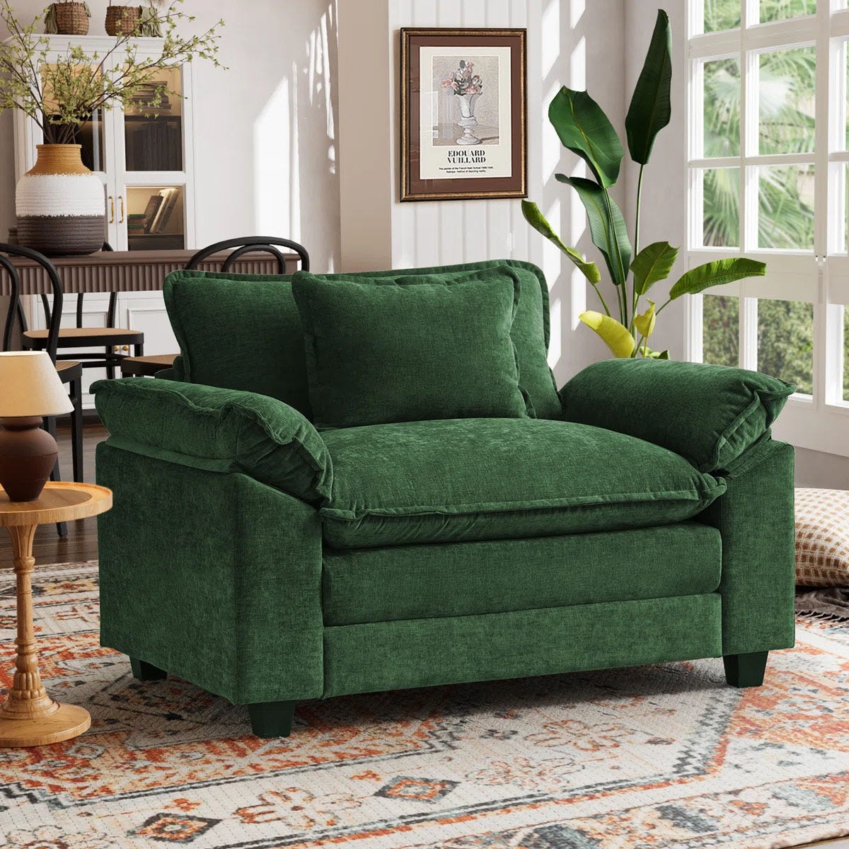 A green velvet loveseat with plush cushions is situated in a living room featuring a patterned rug, wall art, and various decor items, including a potted plant and a side table with a lamp.