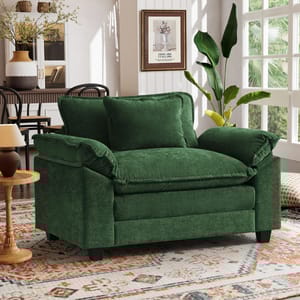 A green velvet loveseat with plush cushions is situated in a living room featuring a patterned rug, wall art, and various decor items, including a potted plant and a side table with a lamp.