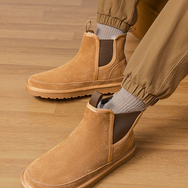 Tan suede ankle boots with elastic side panels and pull tabs, paired with brown cuffed pants and gray socks, displayed on a wood floor.