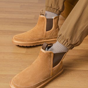Tan suede ankle boots with elastic side panels and pull tabs, paired with brown cuffed pants and gray socks, displayed on a wood floor.
