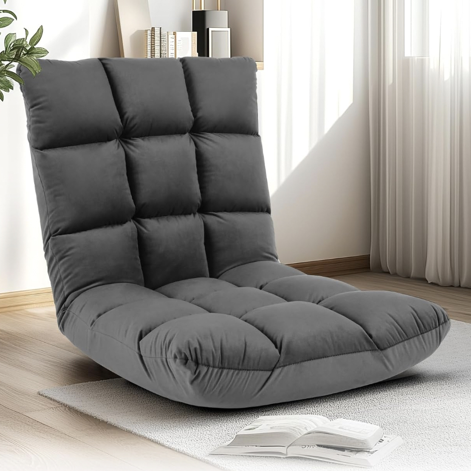 A comfortable, gray, tufted floor chair with a high backrest placed on a light rug in a room with wooden flooring, books, and a plant nearby.