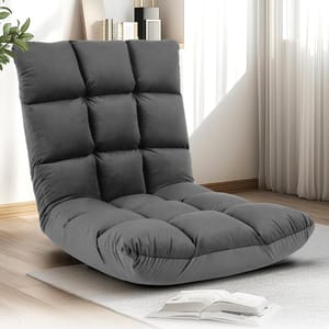 A comfortable, gray, tufted floor chair with a high backrest placed on a light rug in a room with wooden flooring, books, and a plant nearby.