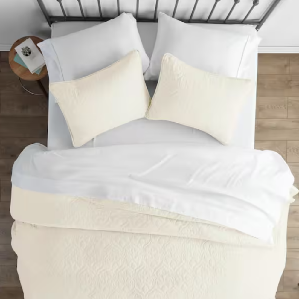 A neatly made bed features a white duvet, matching pillowcases, and two beige accent pillows on a metal headboard. A small wooden side table holds books and decor.