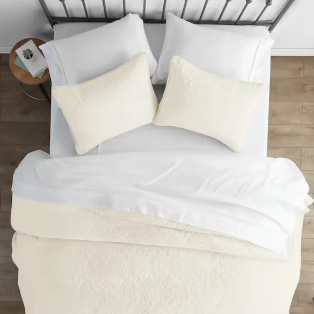 A neatly made bed features a white duvet, matching pillowcases, and two beige accent pillows on a metal headboard. A small wooden side table holds books and decor.