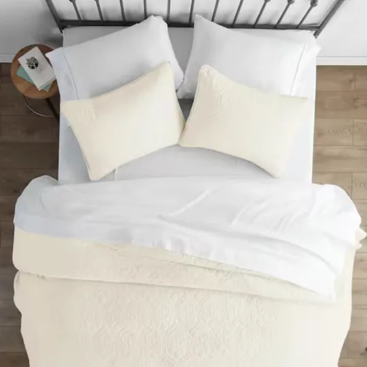 A neatly made bed features a white duvet, matching pillowcases, and two beige accent pillows on a metal headboard. A small wooden side table holds books and decor.