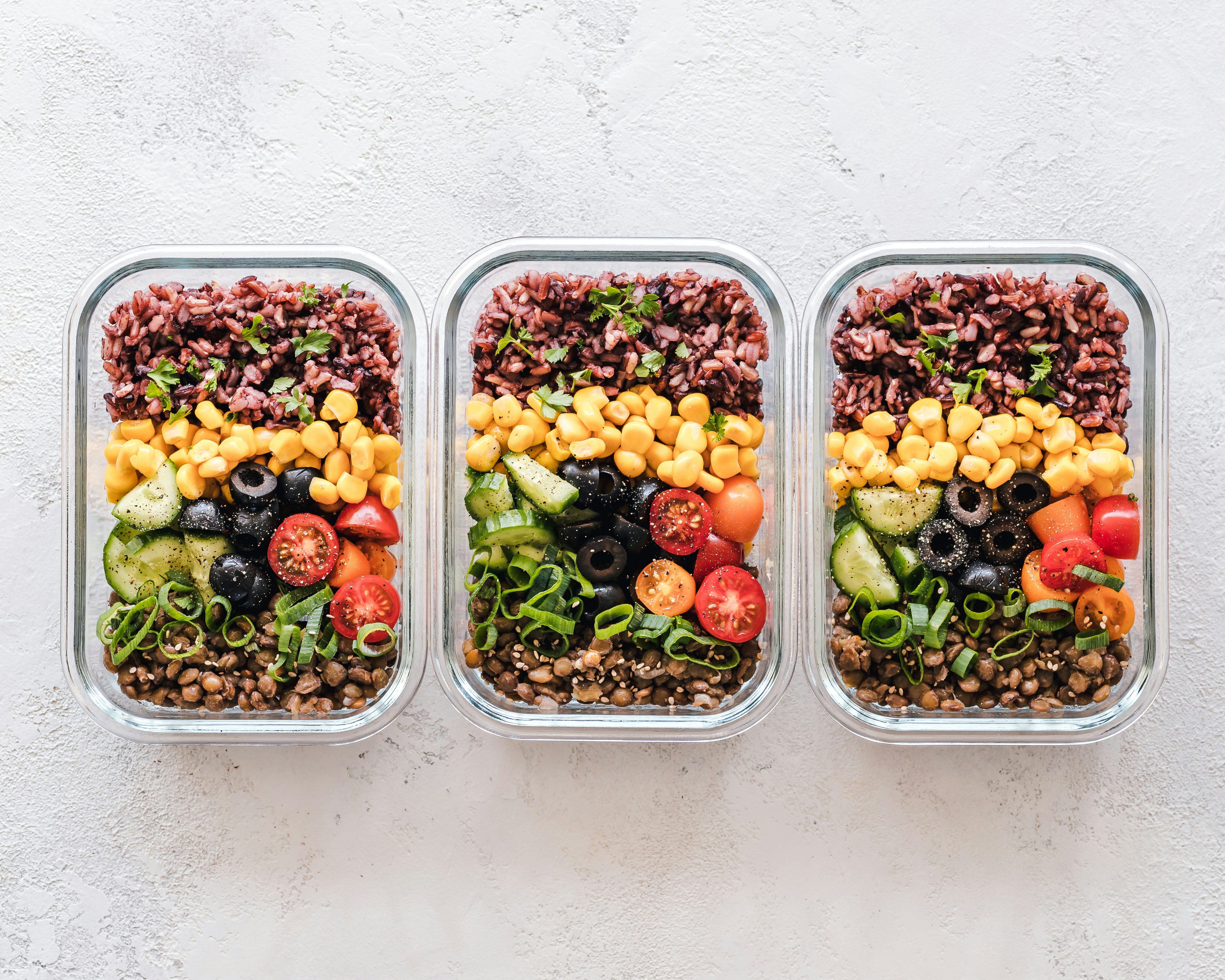 Three meal prep containers with brown rice, lentils, chickpeas, cucumbers, tomatoes, olives, and green onions.