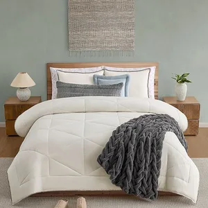 A neatly made bed features a cream quilted comforter and a chunky gray knitted throw, flanked by wooden nightstands with matching lamps and minimal decor.