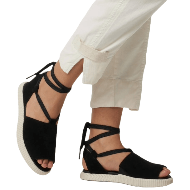 Black strappy sandals with a peep-toe design, featuring ankle wrap ties and white textured soles. Styled with light-colored pants rolled up at the cuffs.