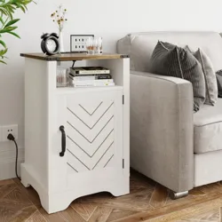 A white side table with a chevron-patterned door is placed next to a beige sofa. It features a wooden top with books, an alarm clock, and glassware on it.