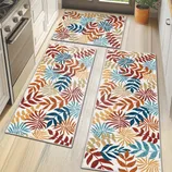 Set of three kitchen floor mats with a tropical leaf pattern in red, orange, blue, and beige tones. They are placed in front of a stove and sink in a bright kitchen with wooden cabinets and a potted plant.