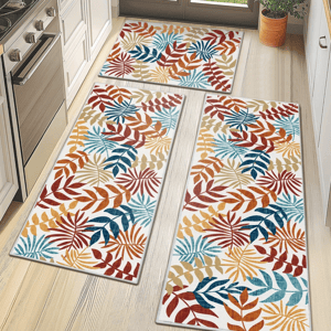 Set of three kitchen floor mats with a tropical leaf pattern in red, orange, blue, and beige tones. They are placed in front of a stove and sink in a bright kitchen with wooden cabinets and a potted plant.