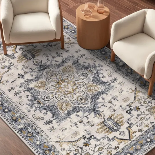 Two cream-colored armchairs are placed around a small round wooden table on a patterned rug with blue, white, and gold tones.
