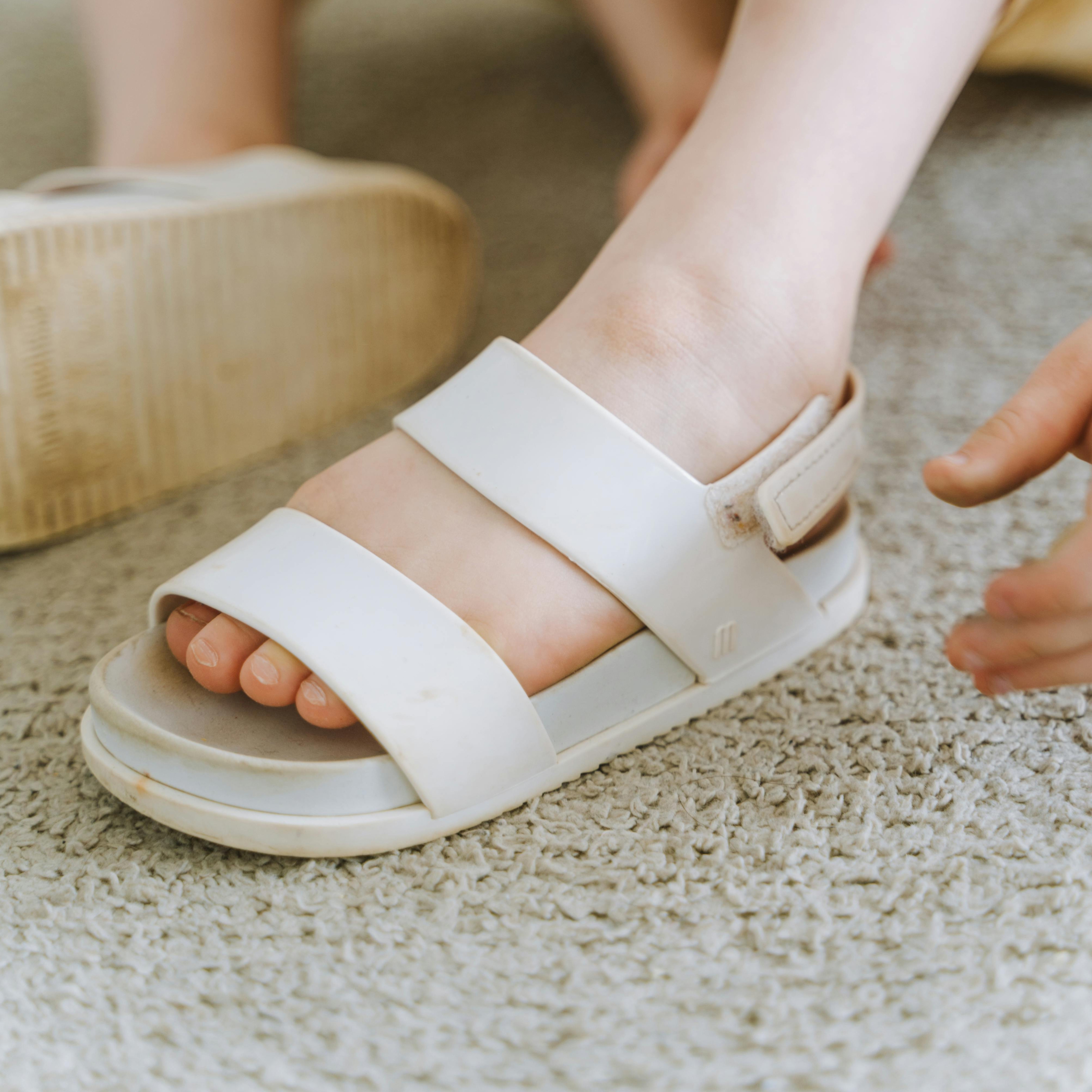 White slide sandals with two straps on a person's foot, against a carpeted floor.