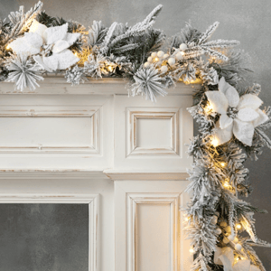 A frosted garland with white poinsettias and warm string lights decorates a white mantel, set against a gray wall.