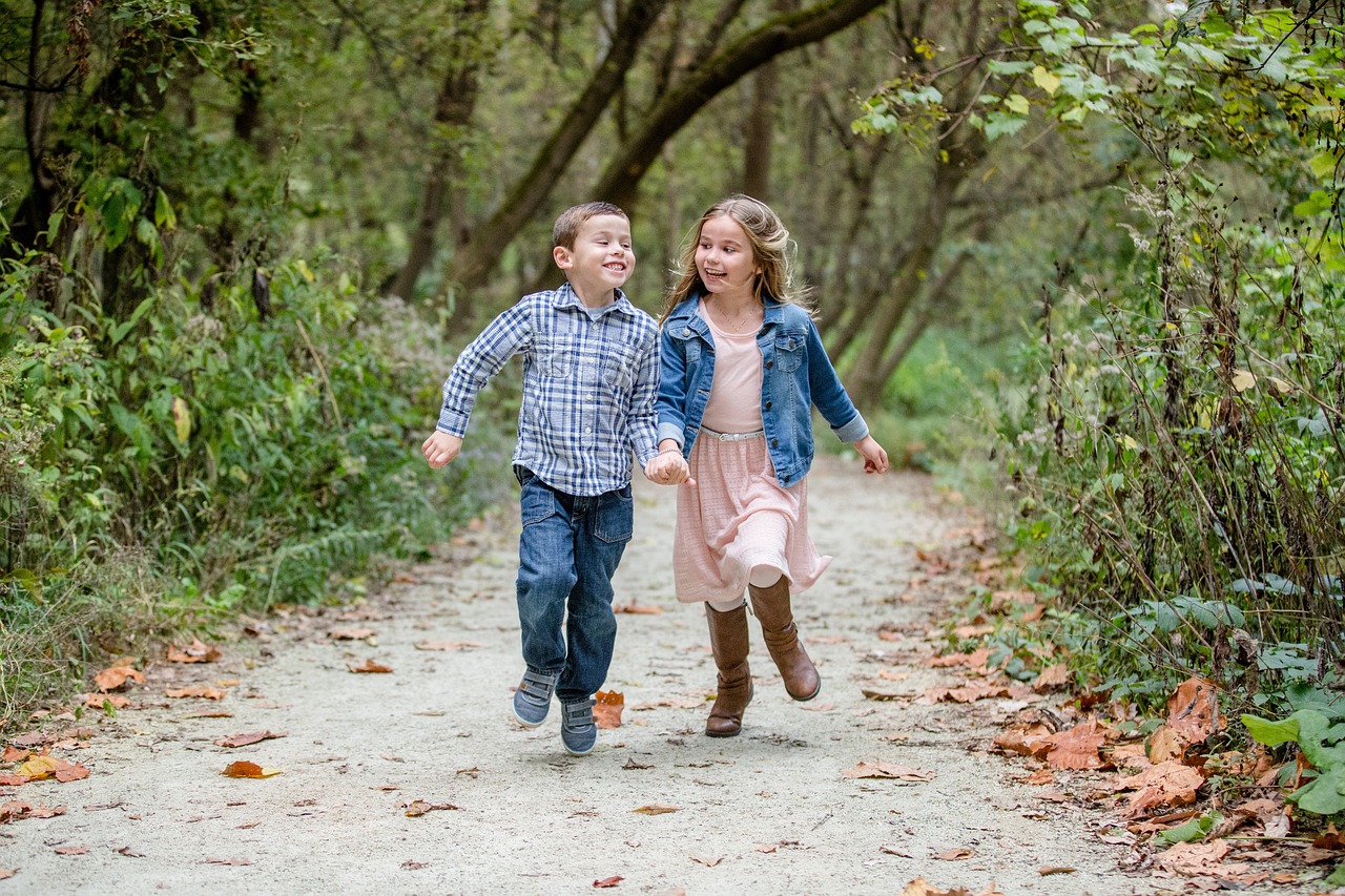 Two children are running on a forest path with leaves scattered around, the boy in a plaid shirt and denim pants, and the girl in a pink skirt with a denim jacket.