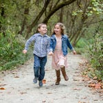 Two children are running on a forest path with leaves scattered around, the boy in a plaid shirt and denim pants, and the girl in a pink skirt with a denim jacket.