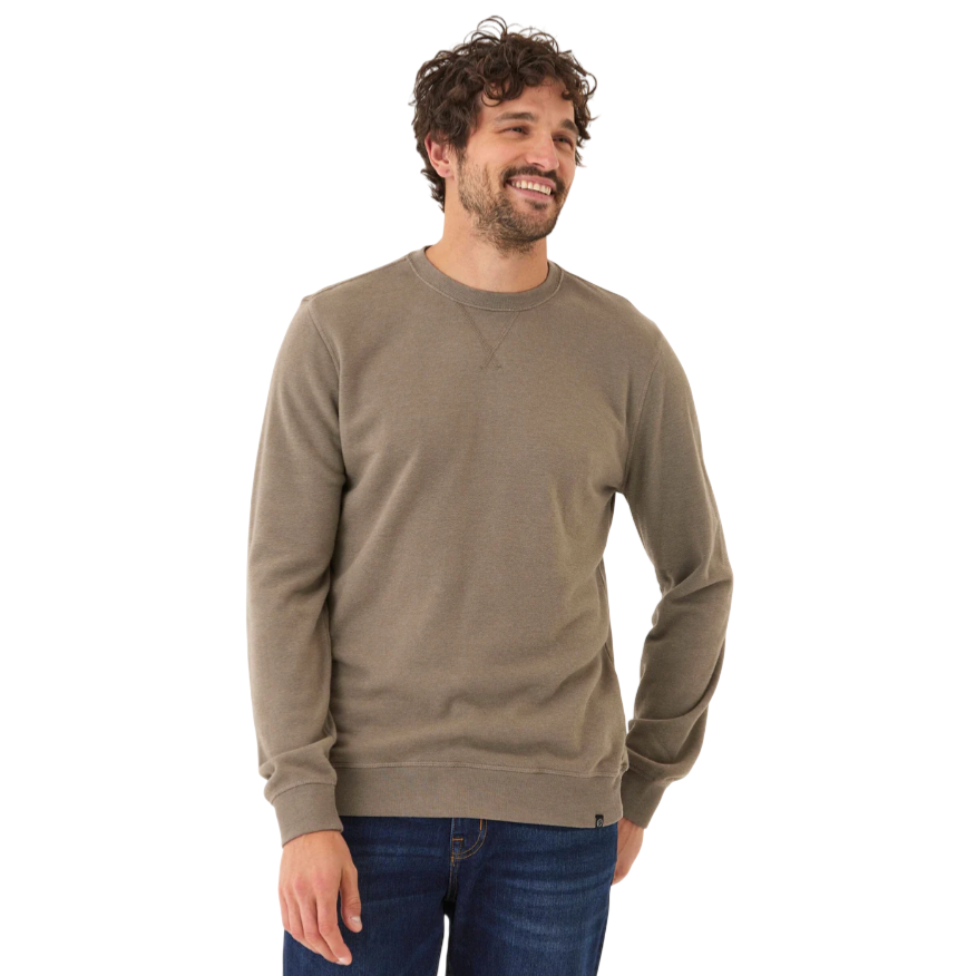 A man is wearing a plain taupe sweatshirt and blue jeans, smiling with his hands at his sides.