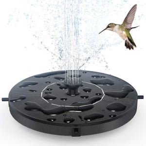 A round, black solar-powered birdbath fountain featuring a central water spout creating vertical jets, and a hummingbird hovering nearby.
