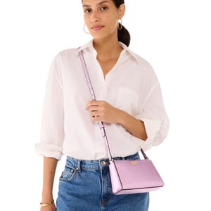 The Kate Spade Mini Crossbody is a small, sleek lavender bag with a long adjustable strap and subtle gold detailing, designed for casual wear. It complements a simple outfit with a white blouse and blue jeans.