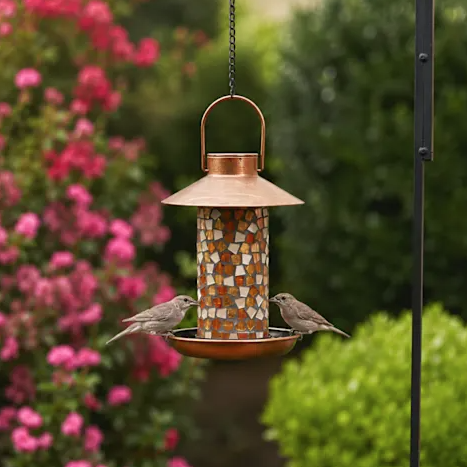 Copper mosaic bird feeder with two birds feeding, set against a garden backdrop featuring vibrant pink flowers and green shrubs.