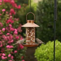 Copper mosaic bird feeder with two birds feeding, set against a garden backdrop featuring vibrant pink flowers and green shrubs.