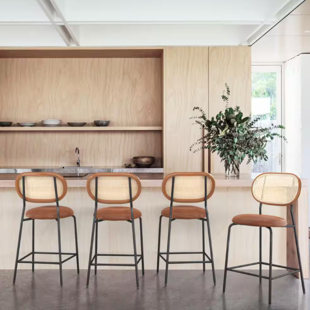 Four counter height bar stools featuring rattan backs, cushioned seats in a warm brown tone, and black metal legs with footrests. They are positioned in front of a light wood bar with minimalist decor.