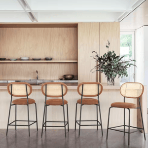 Four counter height bar stools featuring rattan backs, cushioned seats in a warm brown tone, and black metal legs with footrests. They are positioned in front of a light wood bar with minimalist decor.