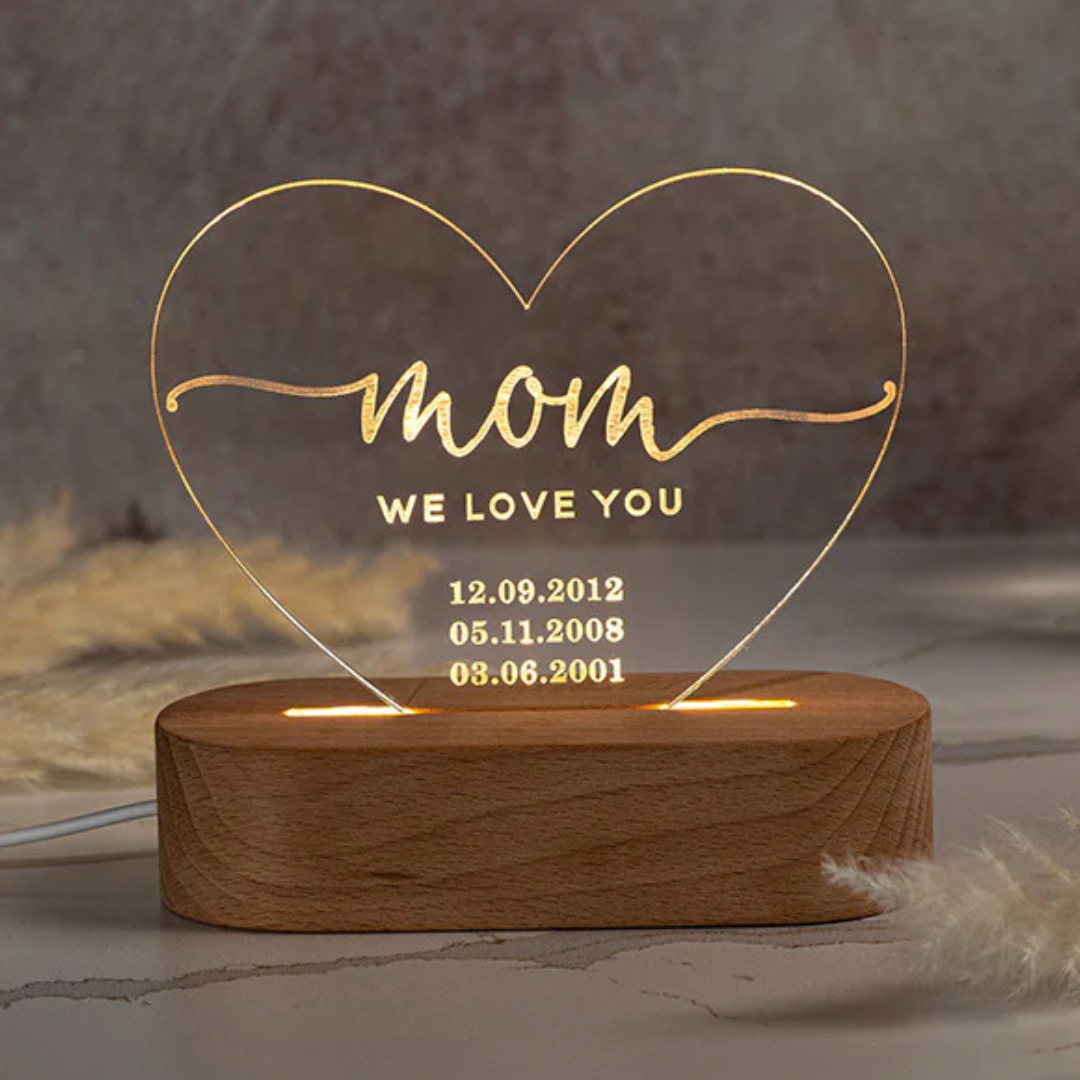 Heart-shaped LED night light with \“mom, we love you\“ text and three date engravings: 12.09.2012, 05.11.2008, 03.06.2001, mounted on a wooden base.