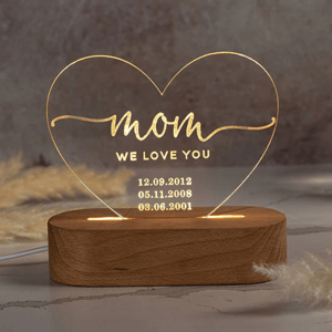 Heart-shaped LED night light with \“mom, we love you\“ text and three date engravings: 12.09.2012, 05.11.2008, 03.06.2001, mounted on a wooden base.