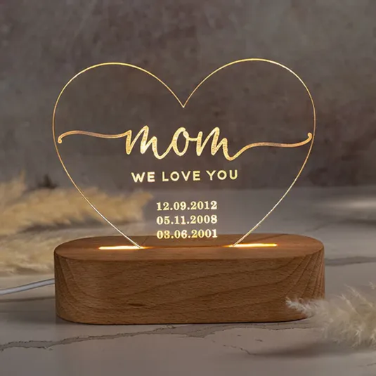Heart-shaped LED night light with \“mom, we love you\“ text and three date engravings: 12.09.2012, 05.11.2008, 03.06.2001, mounted on a wooden base.