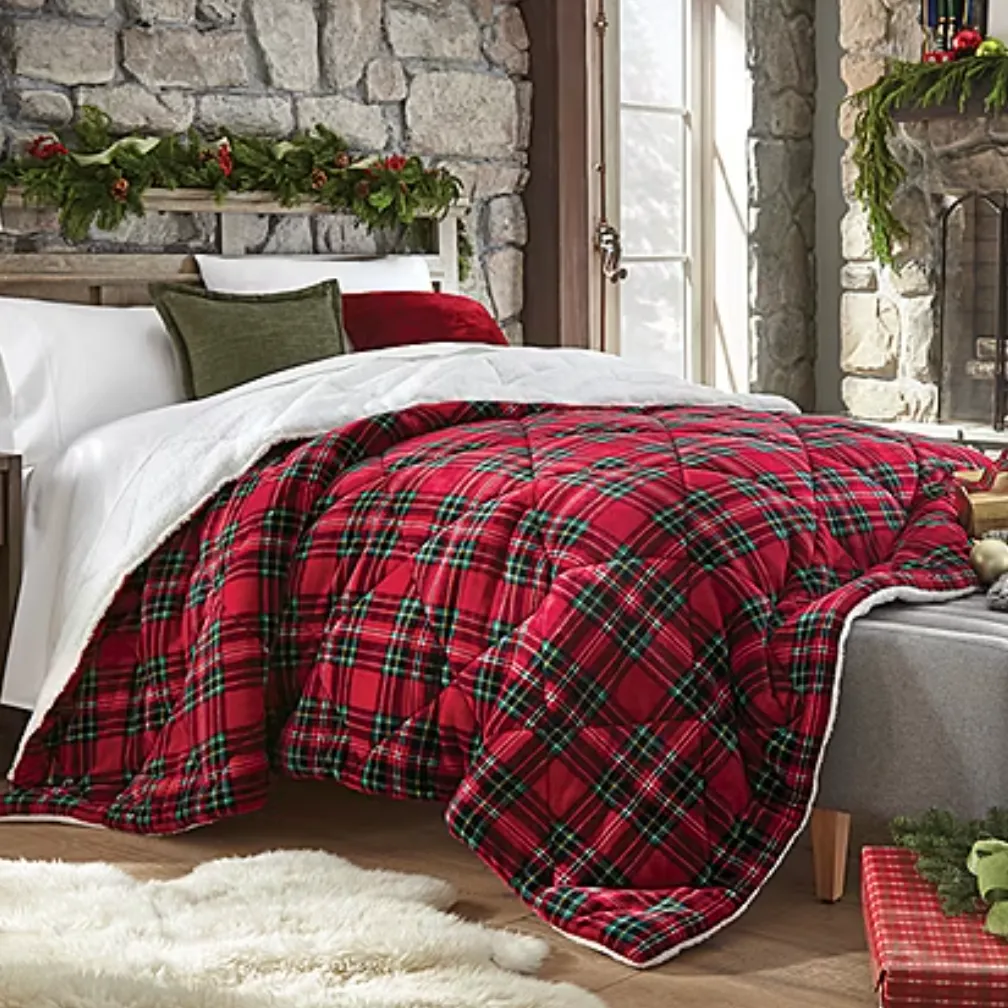 A bed with a red plaid comforter is set with white, green, and red pillows in a cozy room featuring a stone wall, a window, and a fireplace adorned with greenery and holiday decor. A soft, cream rug is placed on the wooden floor.