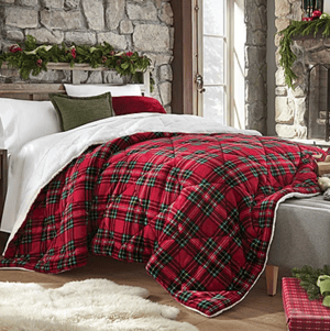 A bed with a red plaid comforter is set with white, green, and red pillows in a cozy room featuring a stone wall, a window, and a fireplace adorned with greenery and holiday decor. A soft, cream rug is placed on the wooden floor.