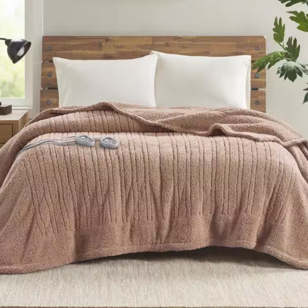 A cozy brown textured electric blanket with remote controls sits on a bed with a wooden headboard and white pillows, accompanied by a bedside lamp and a potted plant.