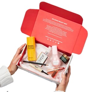 A beauty box with skincare and cosmetic items including Nécessaire body serum, Herbivore facial mist, eye cream, brow pencil, and lip product, all arranged on pink tissue paper inside a red and white box.