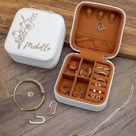 White jewelry box with floral design and name \“Michelle,\“ containing rings, earrings, and a necklace, displayed open on a wooden surface alongside additional jewelry pieces like a bracelet and chain.