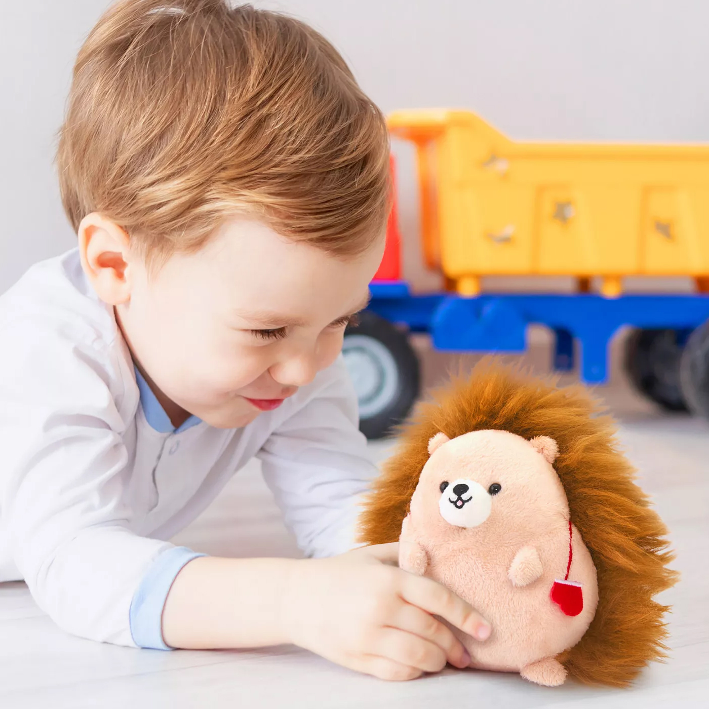 A young child plays with a plush hedgehog toy on the floor. A yellow and blue toy truck is visible in the background.