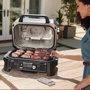 A portable grill with meats cooking on it, and a smartphone showing a temperature app next to the grill.