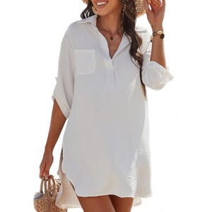 White long-sleeve shirt dress with a collar and chest pocket, styled with rolled-up sleeves. The person is also wearing a brown bracelet and carrying a woven straw handbag.