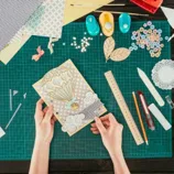A craft workspace with a green cutting mat, paper punches, buttons, a ruler, scissors, pencils, and an intricate hot air balloon card.