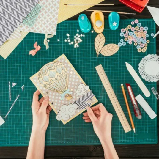 A craft workspace with a green cutting mat, paper punches, buttons, a ruler, scissors, pencils, and an intricate hot air balloon card.