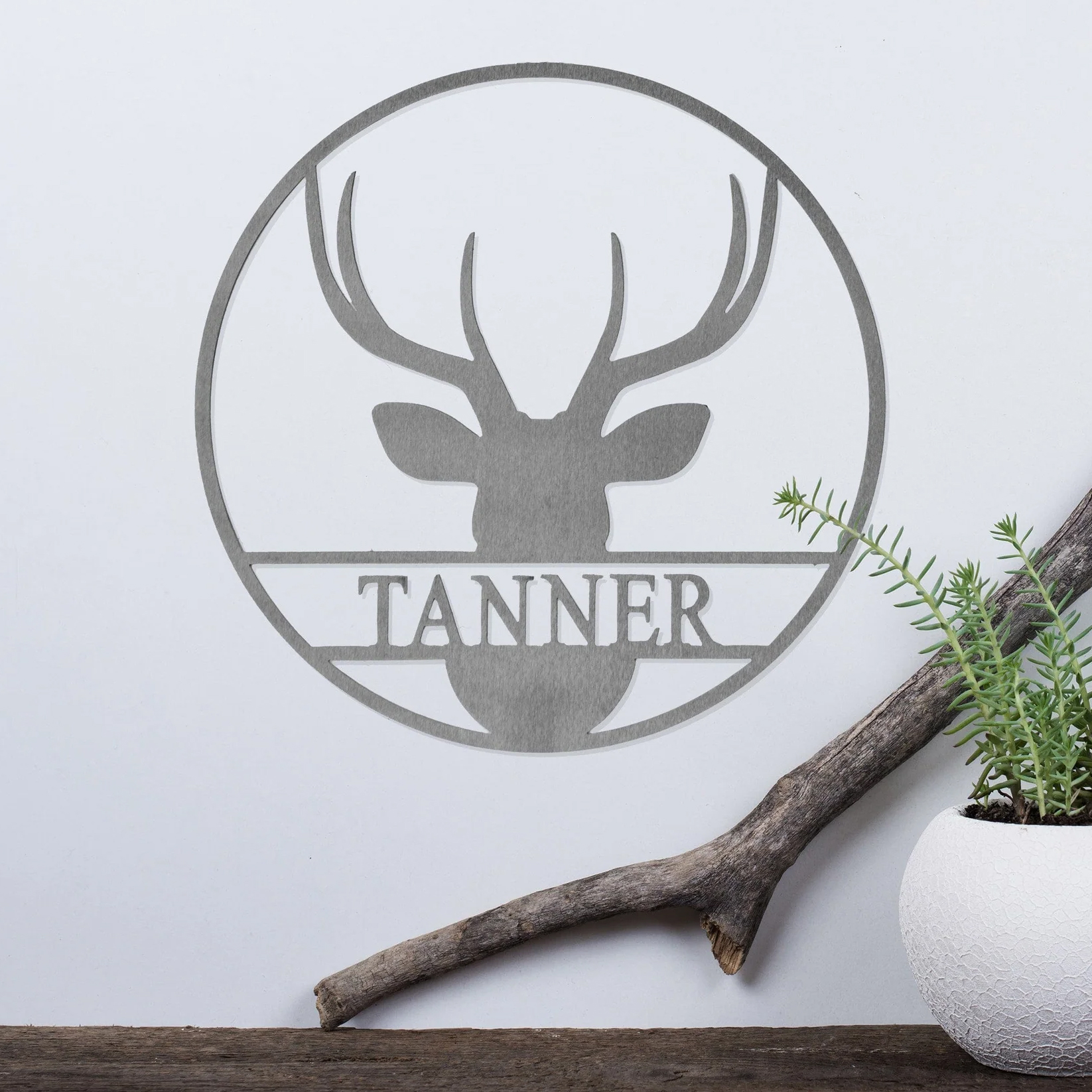 A metal wall art piece features a silhouette of a deer head with antlers, encircled by a ring, and the name \“TANNER\“ beneath the design. It is displayed alongside a white planter and a piece of driftwood.
