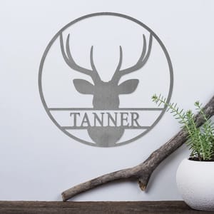 A metal wall art piece features a silhouette of a deer head with antlers, encircled by a ring, and the name \“TANNER\“ beneath the design. It is displayed alongside a white planter and a piece of driftwood.